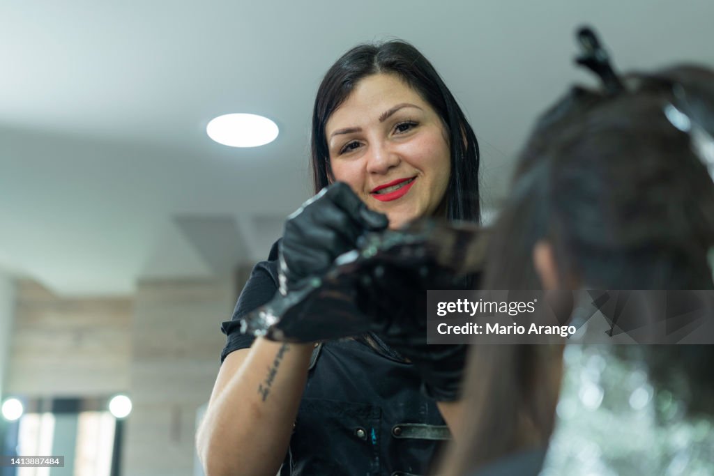Donna proprietaria di un salone di parrucchiere mette la tintura sui capelli del suo cliente mentre guarda la macchina fotografica che la ritrae