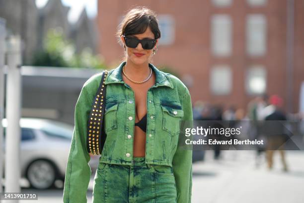 Gili Biegun wears sunglasses, earrings, a necklace, black bras / crop top, a green tie and dye printed denim jacket, a studded bag, matching denim...
