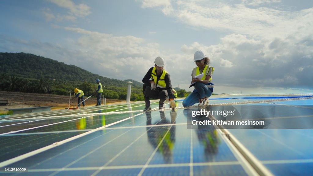 Team contractor, male engineer and female technicians wearing safety uniform, talking about installing plan, check the working system and maintenance solar panel of solar power plant to produce electricity on the roof of factory building.