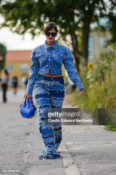 Gili Biegun wears black sunglasses, earrings, a blue denim large cargo / pockets / cropped jacket, bi colored blue / striped ripped / flared denim...