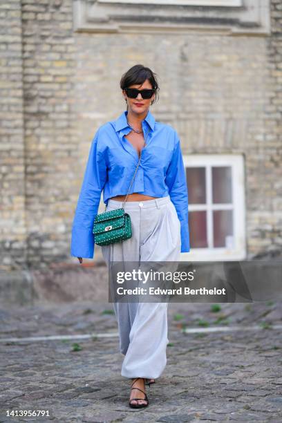 Gili Biegun wears black sunglasses, black earrings, a silver large chain necklace, a blue cropped / oversized shirt, a dark green shiny leather...