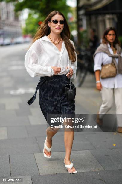 Annabel Rosendahl wears black sunglasses, a gold large chain necklace, a white oversized cotton shirt, black with small white striped print pattern...