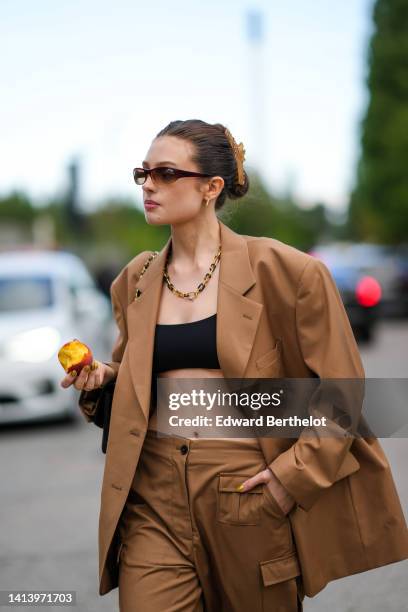 Guest wears brown sunglasses, gold earrings, a gold and black large chain necklace, a beige oversized blazer jacket, matching beige large suit pants,...