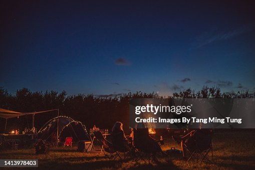 Rear view of a group of people spending time together, sitting in camping chairs  around a burning camp fire in camping field
