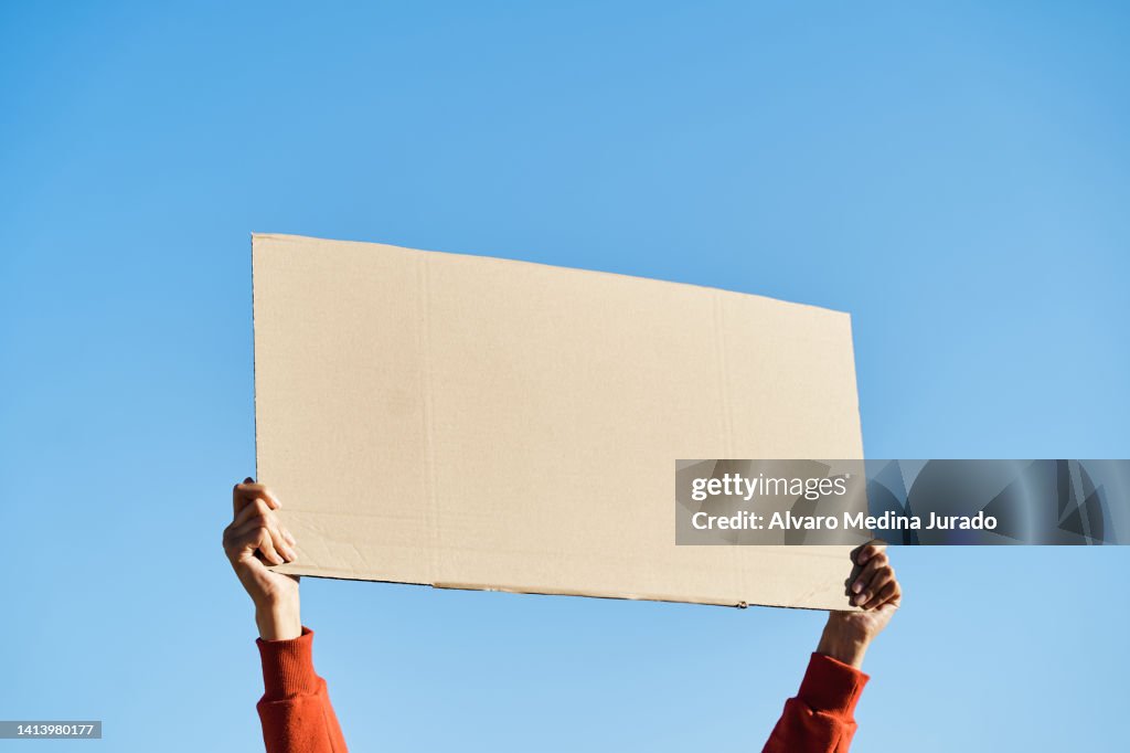 Unrecognizable woman's hands holding a protest banner with no message, with the sky in the background.