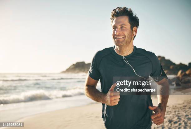 uomo attivo, in forma e sano che fa jogging su una spiaggia mentre ascolta musica in auricolari con splendido cielo limpido all'aperto e spazio per copiare il mare. ragazzo felice, maturo e atletico che corre, fa esercizio di fitness - adulto di mezza età foto e immagini stock