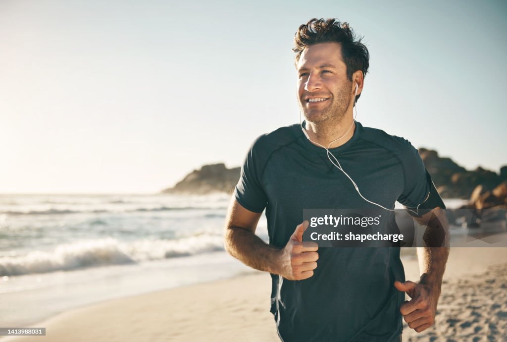 Active, fit and healthy man jogging on a beach while listening to music on earphones with stunning outdoor clear sky and sea copy space. Happy, mature and athletic guy running, doing fitness exercise