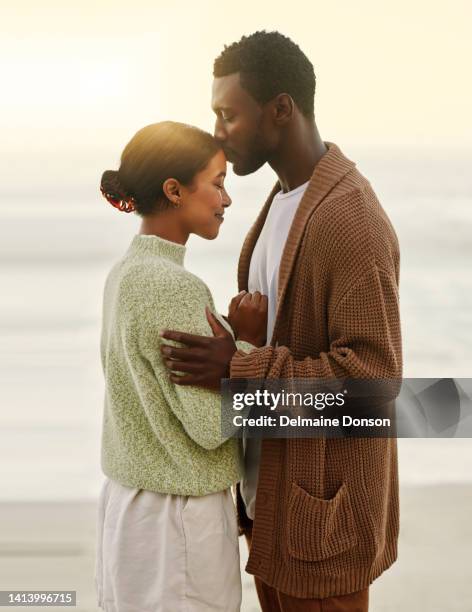 happy, in love and caring couple embracing each other during sunset at the beach. loving african american husband holding his wife and giving her a romantic kiss on her forehead. - black couples kissing stock pictures, royalty-free photos & images