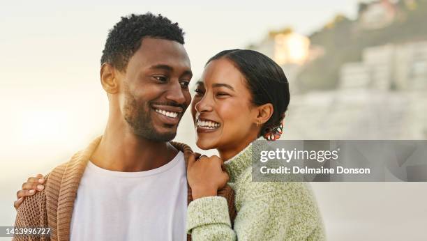 fun, free and happy couple laughing and hugging at the beach, enjoying fresh air and time together. young lovers talking and bonding while being affectionate, sharing a funny joke while walking - air date stockfoto's en -beelden