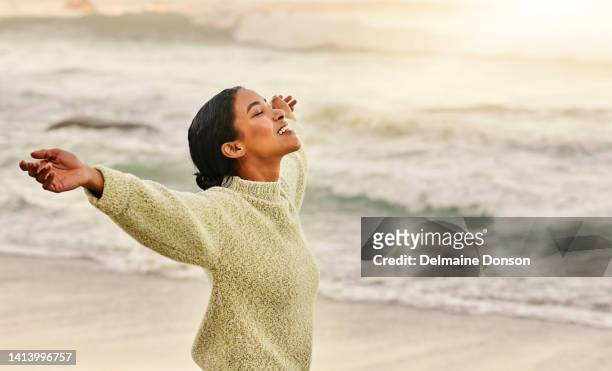 a woman enjoying freedom, being relaxed and carefree on the beach at sunset and looking young, happy and free. smiling black female standing enjoying fresh air and sunshine with ocean in background - african american women in the wind stock pictures, royalty-free photos & images