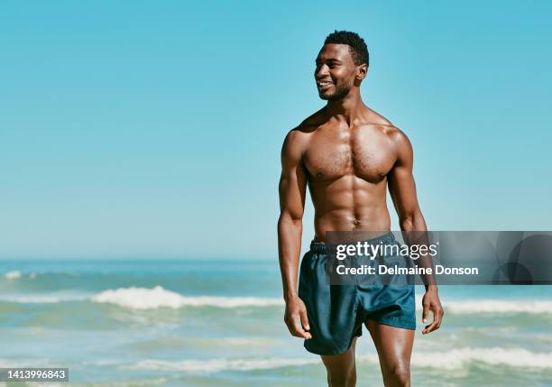 handsome, fit and shirtless man walking on the beach feeling happy, carefree and cheerful while enjoying his summer vacation. black man looking aside and smiling while showing off his hot body - muscle men at beach stockfoto's en -beelden