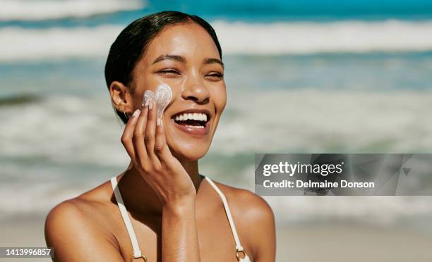 junge frau, die sonnencreme auf ihr gesicht aufträgt, um sich vor uv zu schützen, während sie im urlaub draußen am strand spaß hat. schöne afrikanische frau mit natürlicher und perfekter haut mit ihren händen spf creme - sun on face stock-fotos und bilder