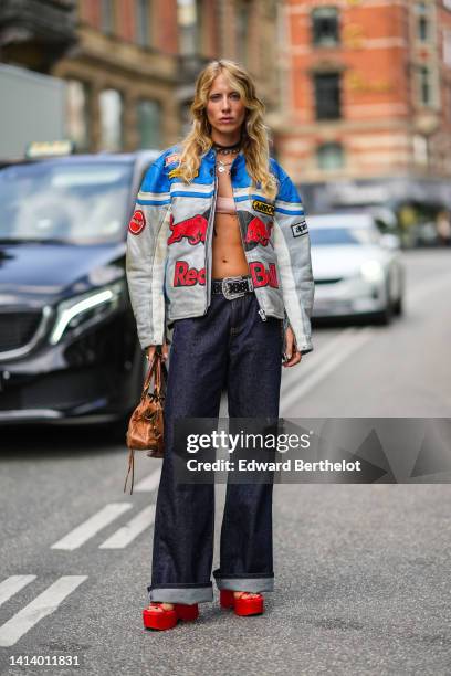 Guest wears earrings, a black leather necklace, silver chain and pendant necklace, a blue / silver / gray / red shiny leather zipper biker jacket...