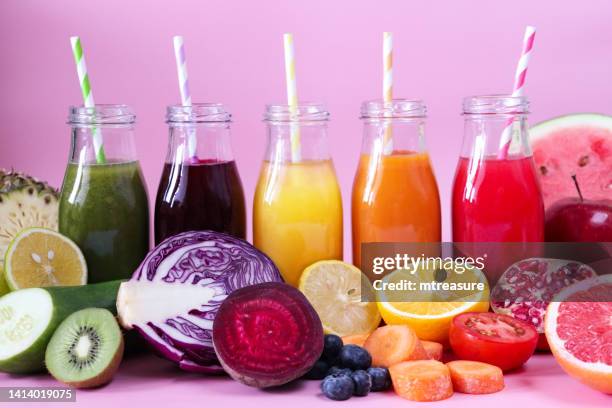 close-up image of row of five glass, screw cap bottles of green, purple, yellow, orange and red fruit and vegetable juice smoothies with stripped drinking straws, fruit and vegetables, pink background, focus on foreground - smoothie stock pictures, royalty-free photos & images