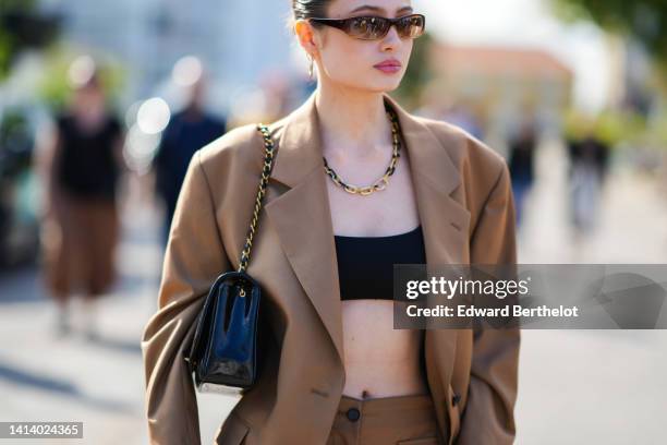 Guest wears brown sunglasses, gold earrings, a gold and black large chain necklace, a beige oversized blazer jacket, matching beige large suit pants,...