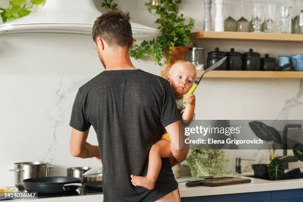 millennial father holds his little son in his arms and cooks breakfast in a bright scandinavian-style kitchen - nanny stock pictures, royalty-free photos & images