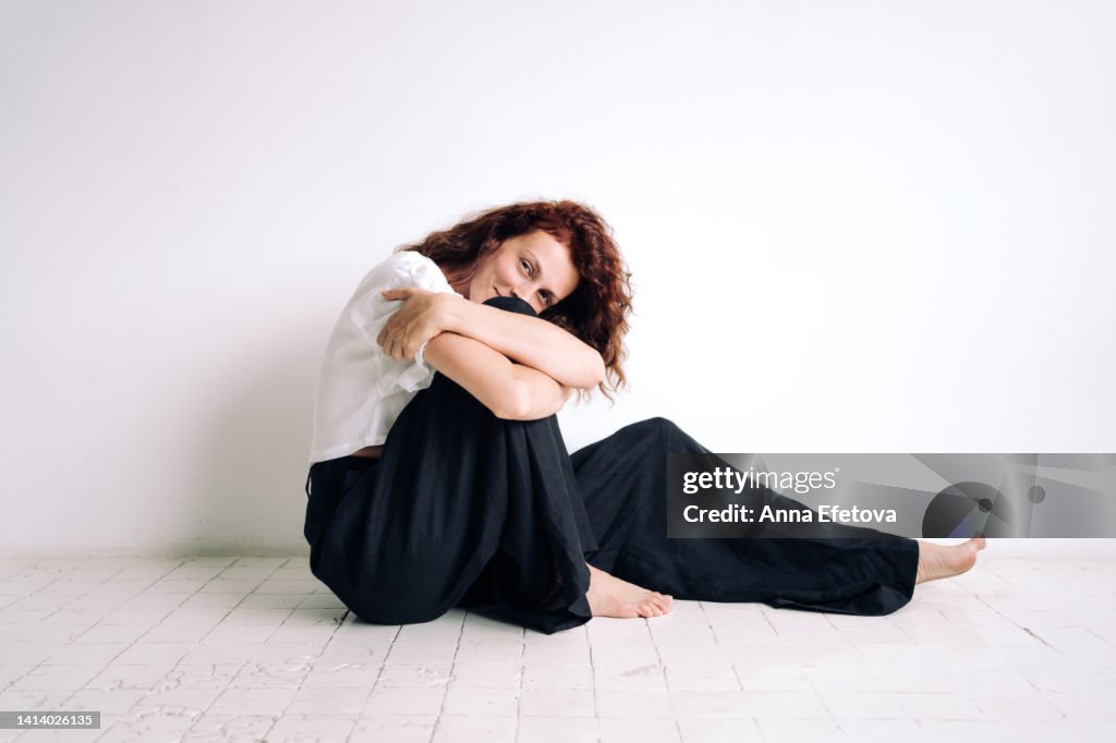 Beautiful authentic young woman with curly brown hair is sitting on the floor hugging her knee against white background. Concept of natural beauty