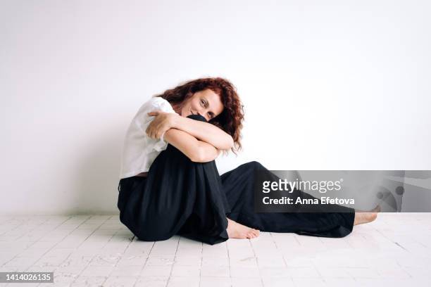 beautiful authentic young woman with curly brown hair is sitting on the floor hugging her knee against white background. concept of natural beauty - krama sig själv bildbanksfoton och bilder