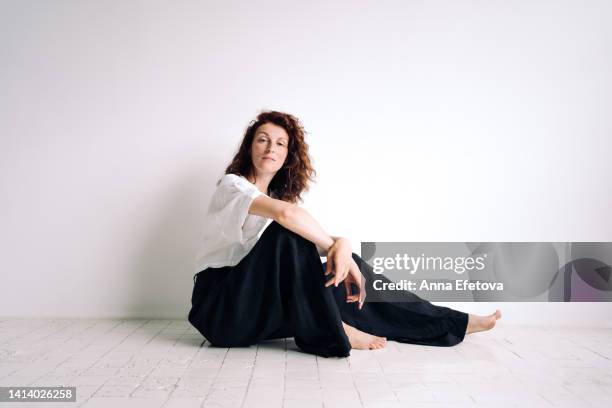 beautiful authentic young woman with curly brown hair is sitting on the floor hugging her knee against white background. concept of natural beauty - hugging knees stock pictures, royalty-free photos & images