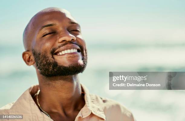 homme africain heureux souriant debout à la plage profitant de la détente et de la chaleur du soleil par une chaude journée d’été. portrait en gros plan d’un homme beau, libre et joyeux avec la mer en arrière-plan - black man beach chair photos et images de collection