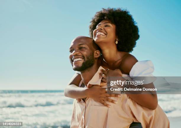 feliz, despreocupada y enamorada pareja en la playa disfrutando de unas relajantes vacaciones de verano al aire libre con olas de mar y cielo azul copiando el fondo del espacio. hombre joven, amoroso y juguetón que le da a la mujer un piggyback - afro fotografías e imágenes de stock