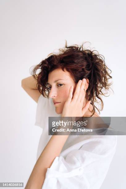 portrait of beautiful authentic young woman with curly brown hair and brown eyes. she is fixing her hairstyle against white wall. concept of natural beauty - curly brown hair stock pictures, royalty-free photos & images