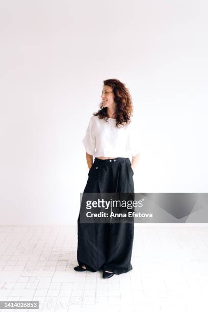 portrait of laughing beautiful authentic young woman with curly brown hair and brown eyes standing against white background with hands in pants pockets. she is wearing loose linen casual clothing. concept of natural beauty - white trousers stockfoto's en -beelden