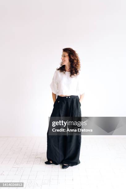 portrait of laughing beautiful authentic young woman with curly brown hair and brown eyes standing against white background with hands in pants pockets. she is wearing loose linen casual clothing. concept of natural beauty - white pants bildbanksfoton och bilder