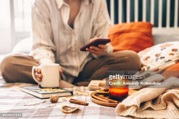 woman sitting on bed in lotus pose using phone and drinking morrning beverage with burning candle - cosy autumn stock pictures, royalty-free photos & images