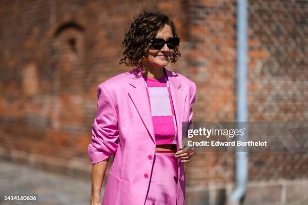 Renata Radzyk wears black sunglasses, a white and neon pink large checkered print pattern cropped top, a pink shiny oversized / long blazer jacket, a...