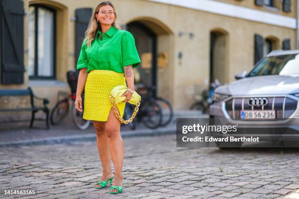 Janka Polliani wears cat eyes glasses, gold earrings, a neon green short sleeves / oversized shirt, a yellow fishnet / mesh slit short skirt, a...