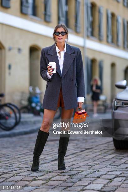 Darja Barannik wears black cat eyes sunglasses, gold earrings, a gold large chain necklace, a white shirt, a dark gray long blazer jacket, camel...