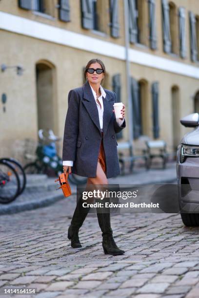 Darja Barannik wears black cat eyes sunglasses, gold earrings, a gold large chain necklace, a white shirt, a dark gray long blazer jacket, camel...