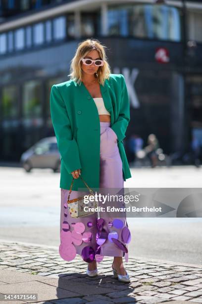 Guest wears pale pink square sunglasses, a gold necklace, a green oversized / buttoned / shoulder pads blazer jacket, pale purple flared pants with...