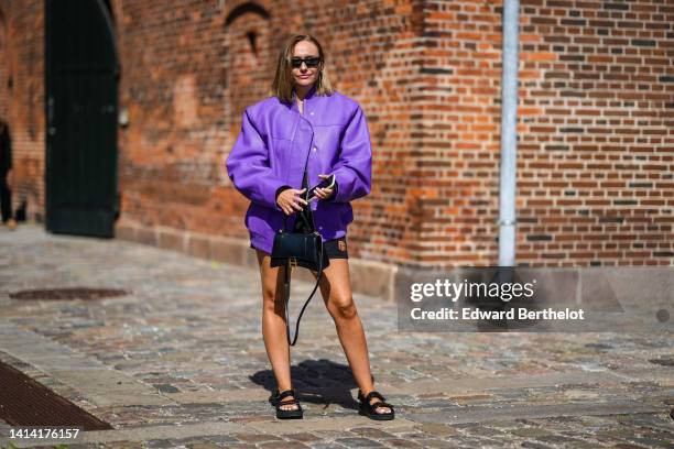 Guest wears black square sunglasses, a white ribbed t-shirt, a purple shiny leather oversized bomber coat, black nylon shorts from Prada, a black...