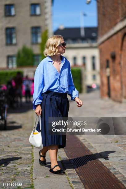 Guest wears black sunglasses, a baby blue with small white striped print pattern shirt, a high waist navy blue pleated midi skirt, a white shiny...