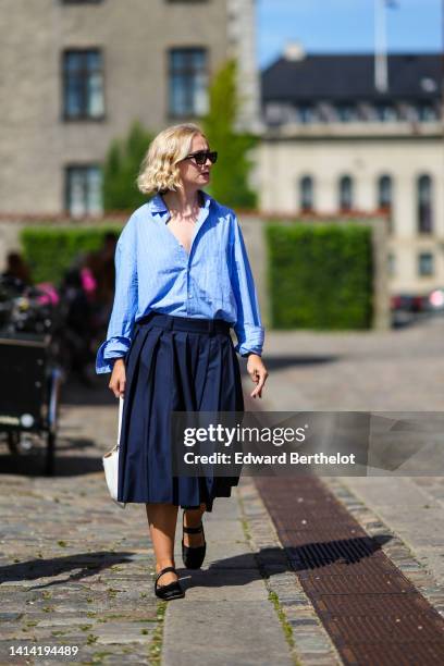 Guest wears black sunglasses, a baby blue with small white striped print pattern shirt, a high waist navy blue pleated midi skirt, a white shiny...