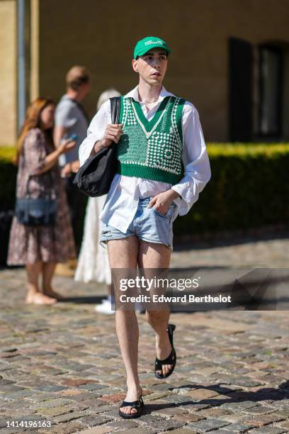 Guest wears a green with white embroidered inscriptions pattern cap, a silver chain necklace, a white shirt, a green and white braided wool /...