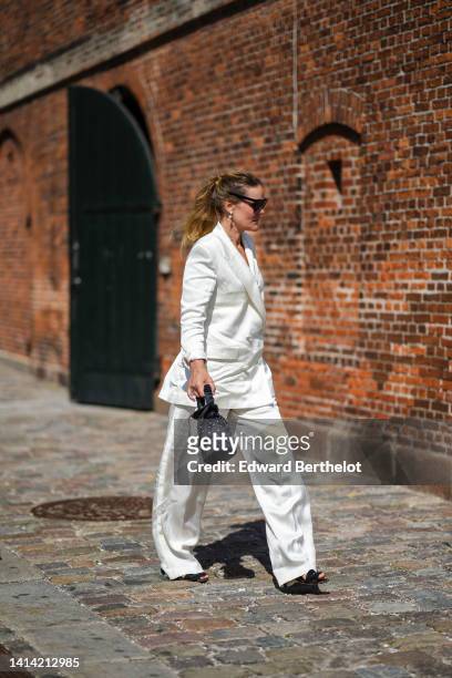 Guest wears black sunglasses, a white pearls pendant earrings, a white long blazer jacket, white large linen pants, a black satin with embroidered...