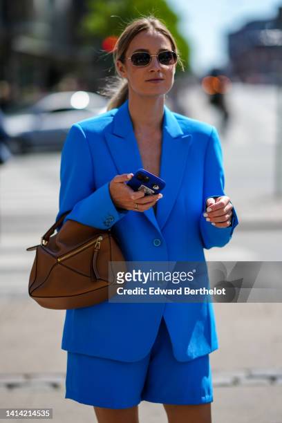 Guest wears sunglasses, a flashy blue blazer jacket, matching flashy blue suit shorts, a brown shiny leather Puzzle handbag from Loewe, rings,...