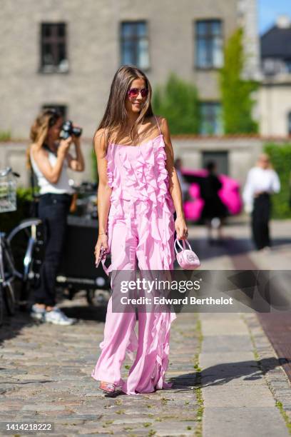 Guest wears pink sunglasses, a pale pink ruffled / fringed jumpsuit, a pale pink shiny satin with embroidered "Ciao Bella" slogan handbag, rings,...