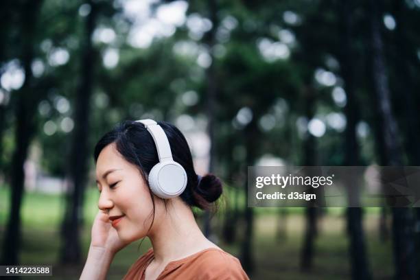 close up of relaxed young asian woman with eyes closed listening to music with headphones in nature, with trees around her outdoors. music, lifestyle and technology - holy city park bildbanksfoton och bilder