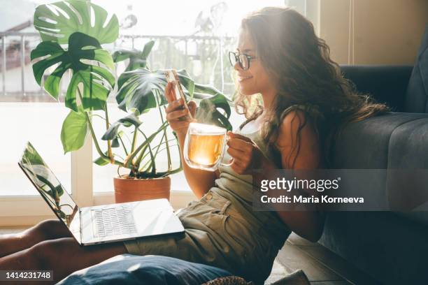 young woman with laptop and phone sitting in living room and drinking tea. - learning resources for reading stock-fotos und bilder