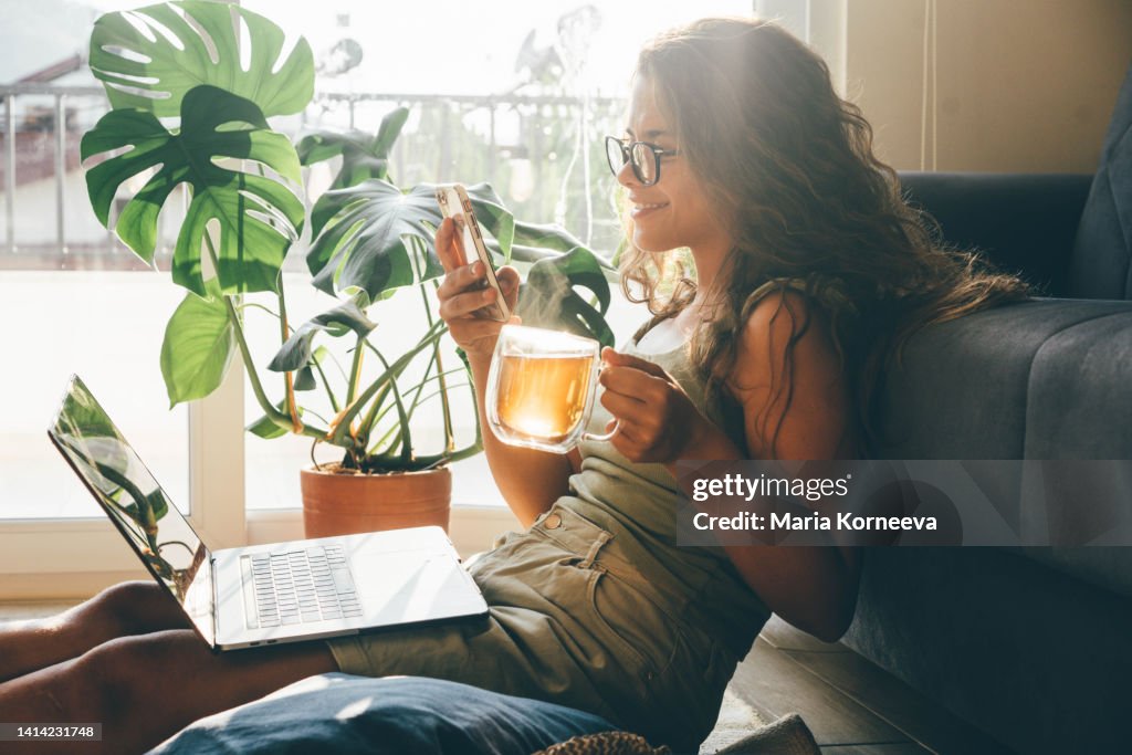 Young woman with laptop and phone sitting in living room and drinking tea.