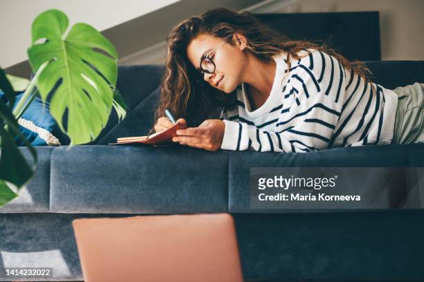 young woman with laptop and notebook lies on the sofa in living room. - learning resources for reading stock-fotos und bilder
