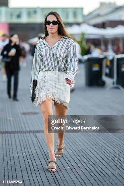 Guest wears black sunglasses, a gray and beige striped print pattern cotton shirt, a brown LV monogram print pattern in coated canvas clutch from...