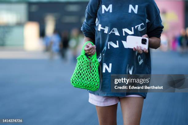 Guest wears a gray with painted white inscription pattern sweater, a neon green mesh / fishnet handbag, pale pink shorts, outside Ganni, during...