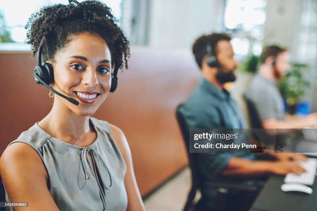 African American female sales team leader or customer service agent working in a call center, talking to a client with a headset. Happy business woman office with diverse colleagues in background.