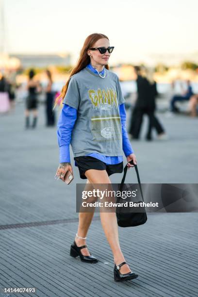 Guest wears black cat eyes sunglasses, a blue shirt, a gray with yellow print pattern t-shirt, black shorts, a black handbag, black shiny varnished...