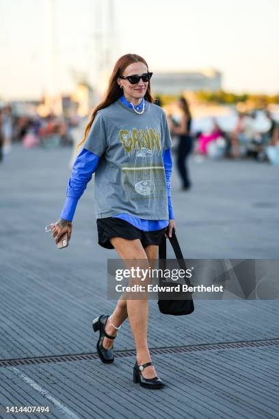 Guest wears black cat eyes sunglasses, a blue shirt, a gray with yellow print pattern t-shirt, black shorts, a black handbag, black shiny varnished...